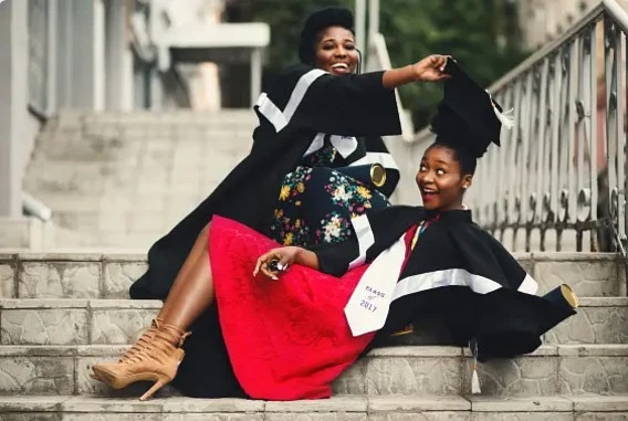 Two students sit on a flight of outdoor stairs in graduation regalia smiling as one takes the other's graduation cap off